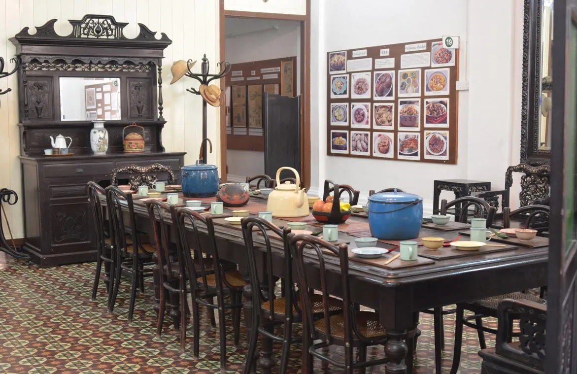 Long dining table with dark wooden chairs, teapots and a carved sideboard on a patterned tile floor