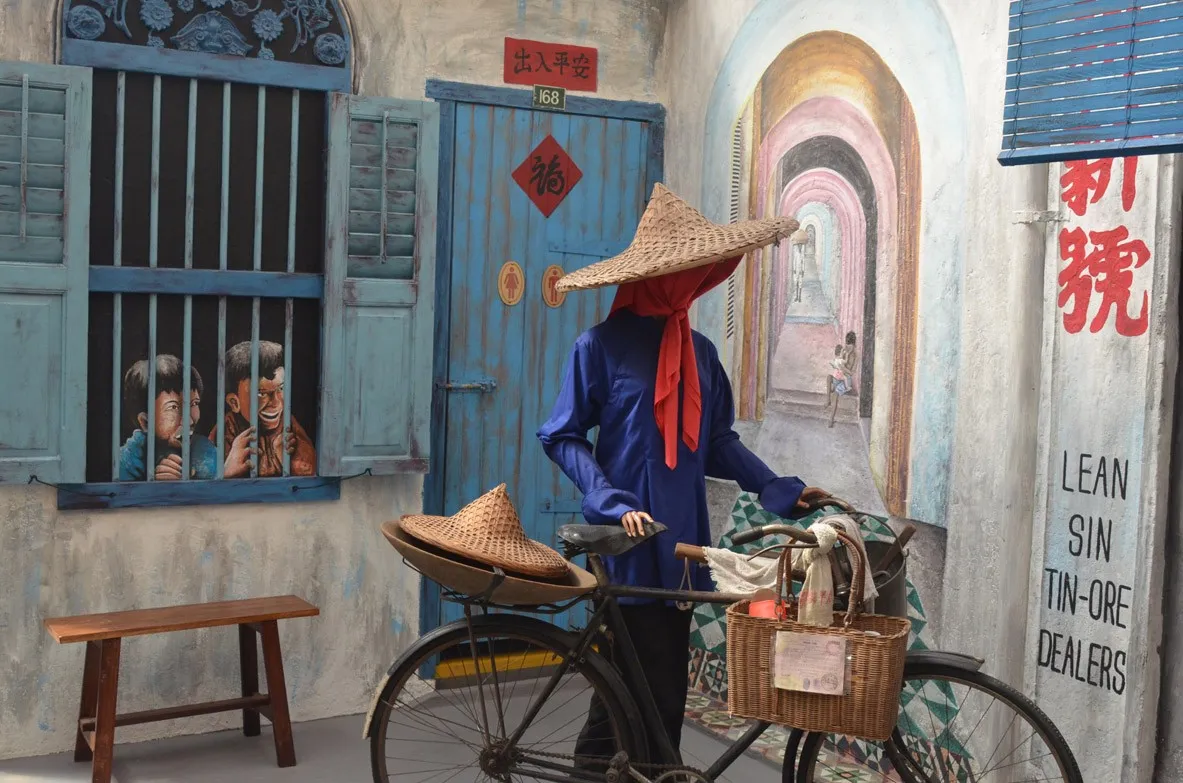 Museum display of a figure in a wide straw hat and red head scarf beside a bicycle loaded with a dulang pan and baskets
