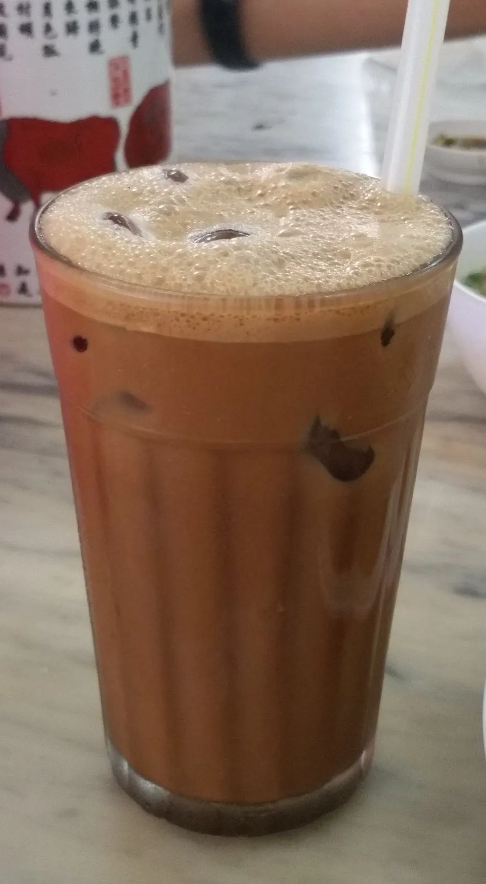 Tall glass of iced Ipoh white coffee with foam, ice and a straw on a marble table