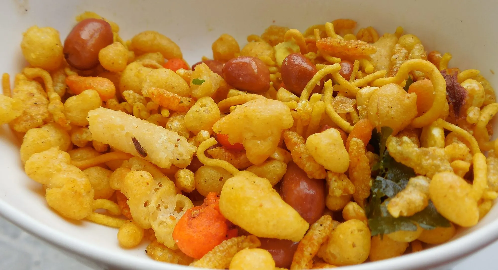 Close-up of a snack mix of peanuts, fried gram-flour pieces and curry leaves in a white bowl