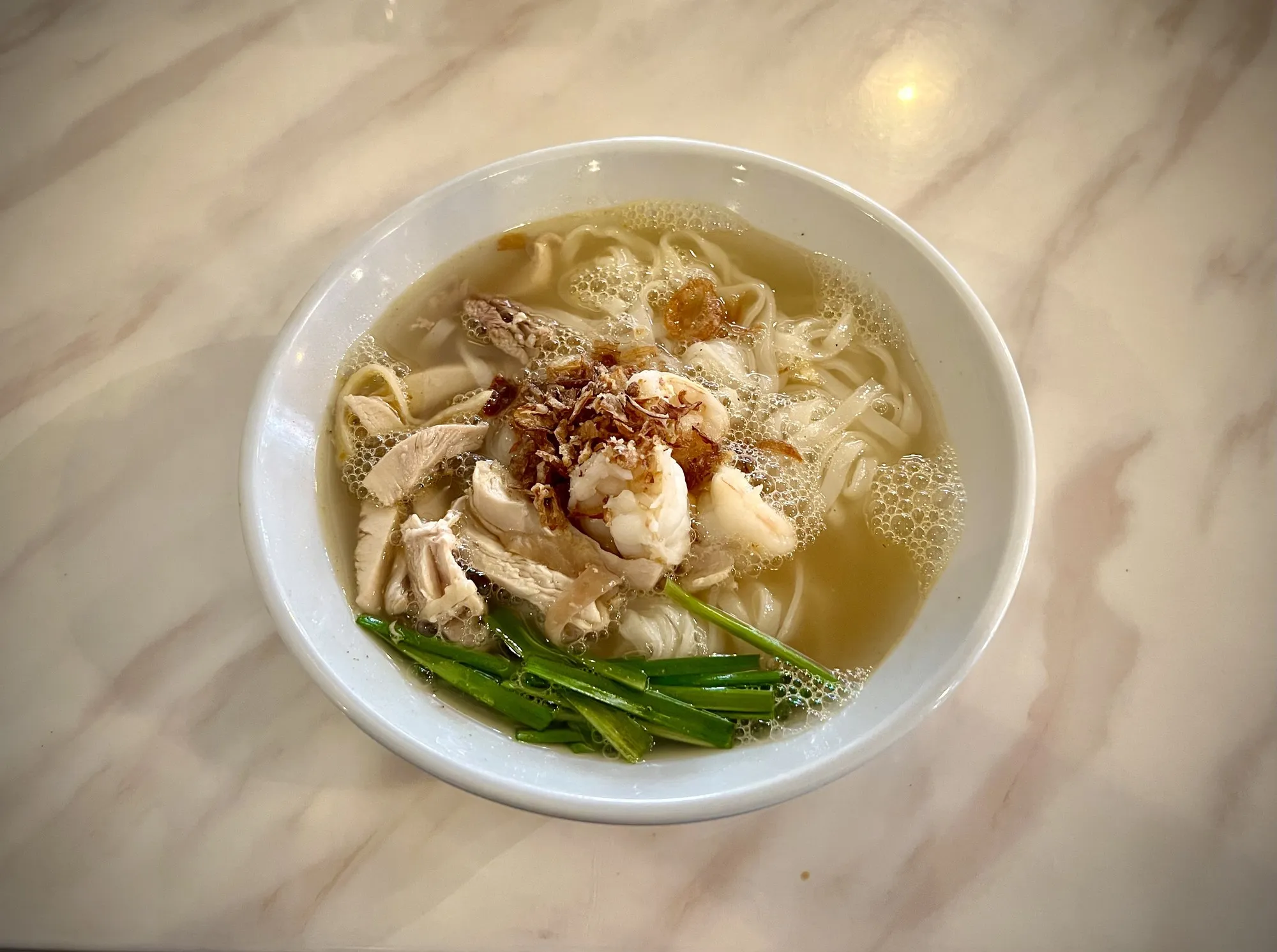 Bowl of kai si hor fun with shredded chicken and chives in clear broth on a marble table