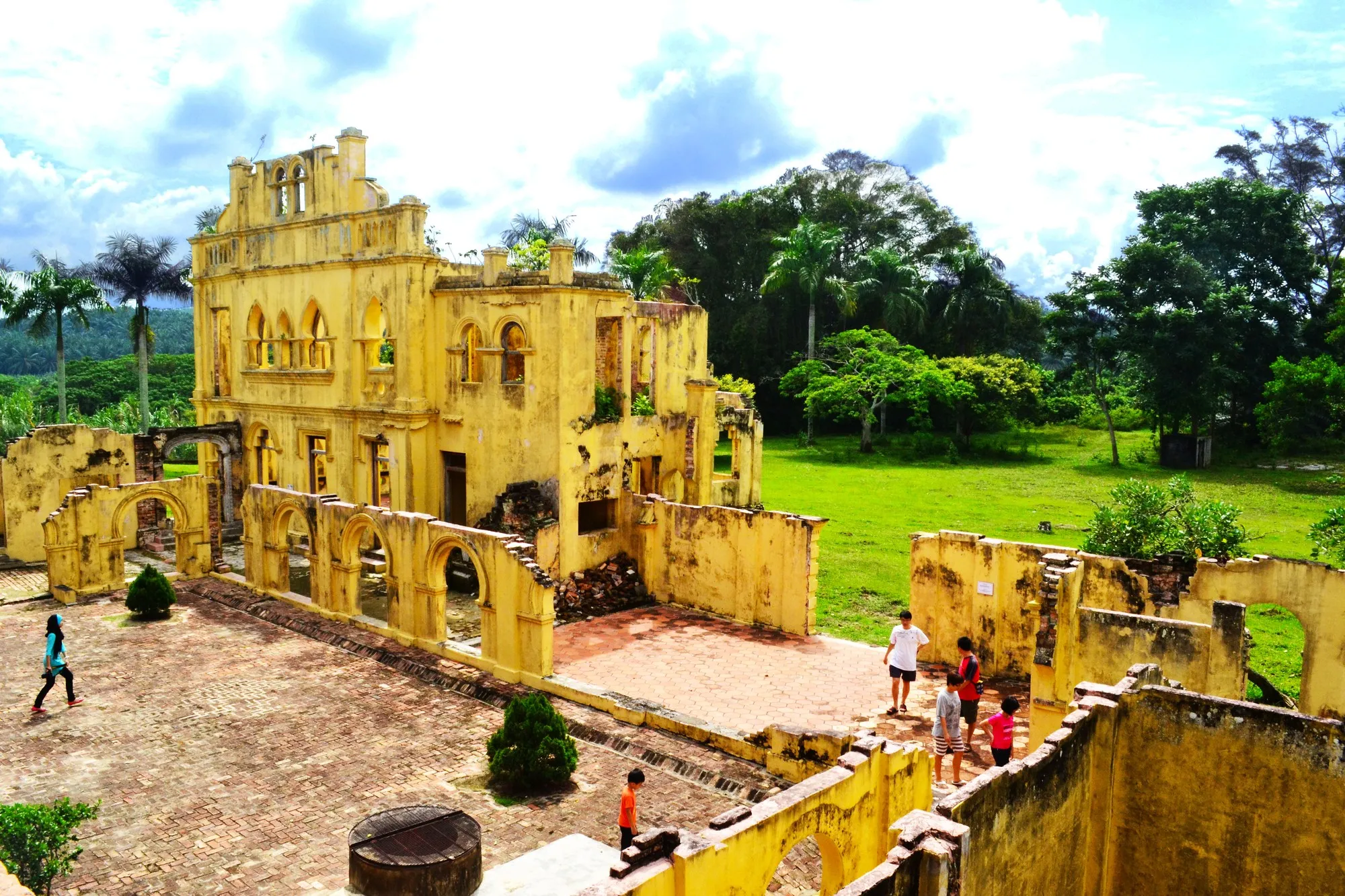 Roofless yellow-plastered ruins of an earlier mansion with arched walls, seen from above beside a lawn