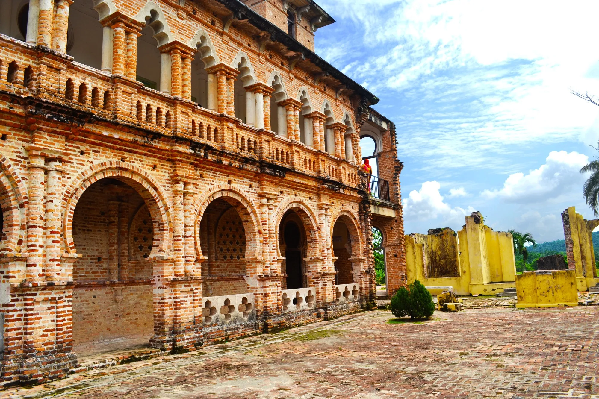 Close view of a weathered brick facade with rows of rounded arches and a balustrade, beside a paved courtyard