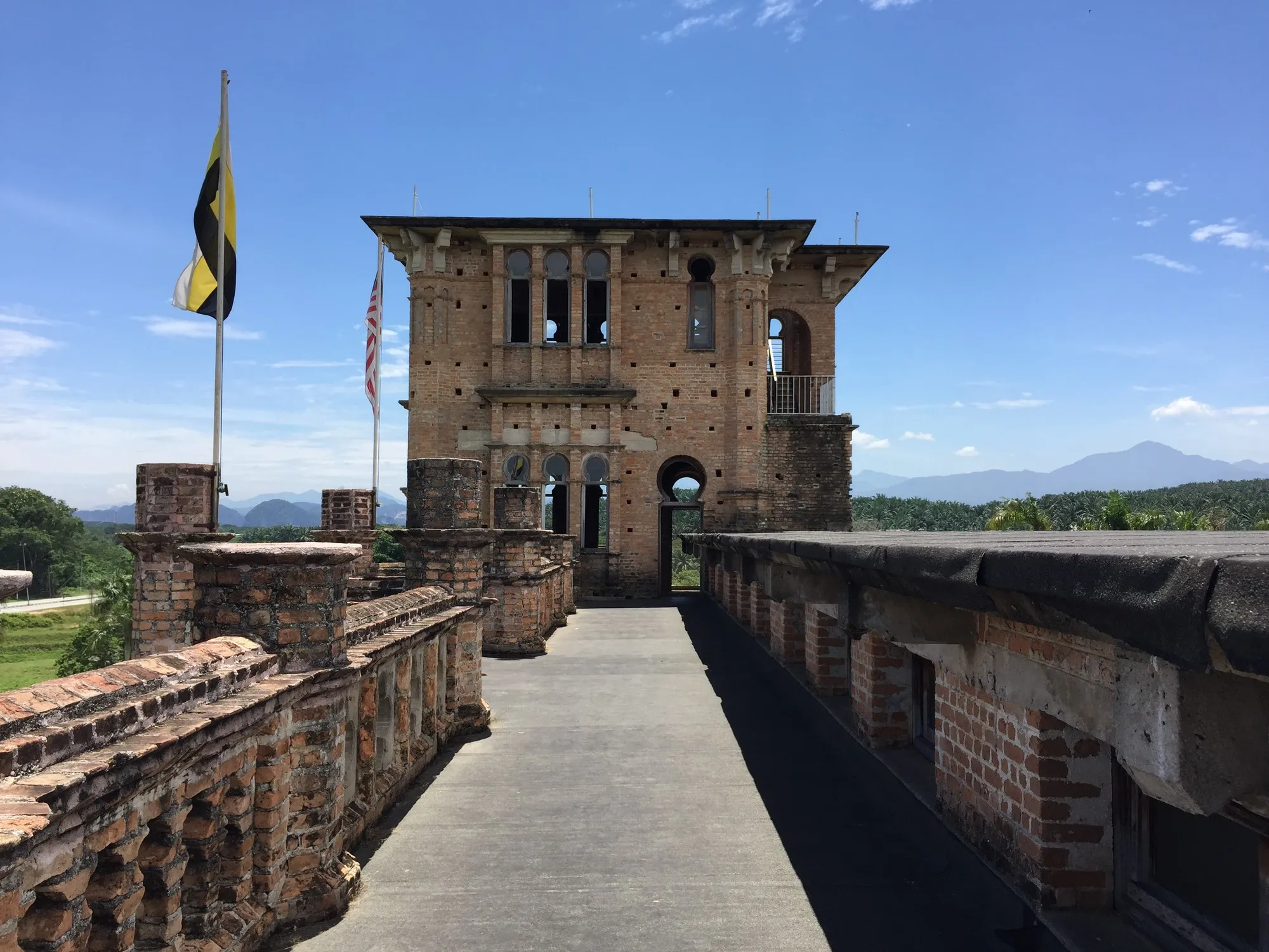 Rooftop walkway with brick parapets leading to a square brick tower, flags and distant hills beyond