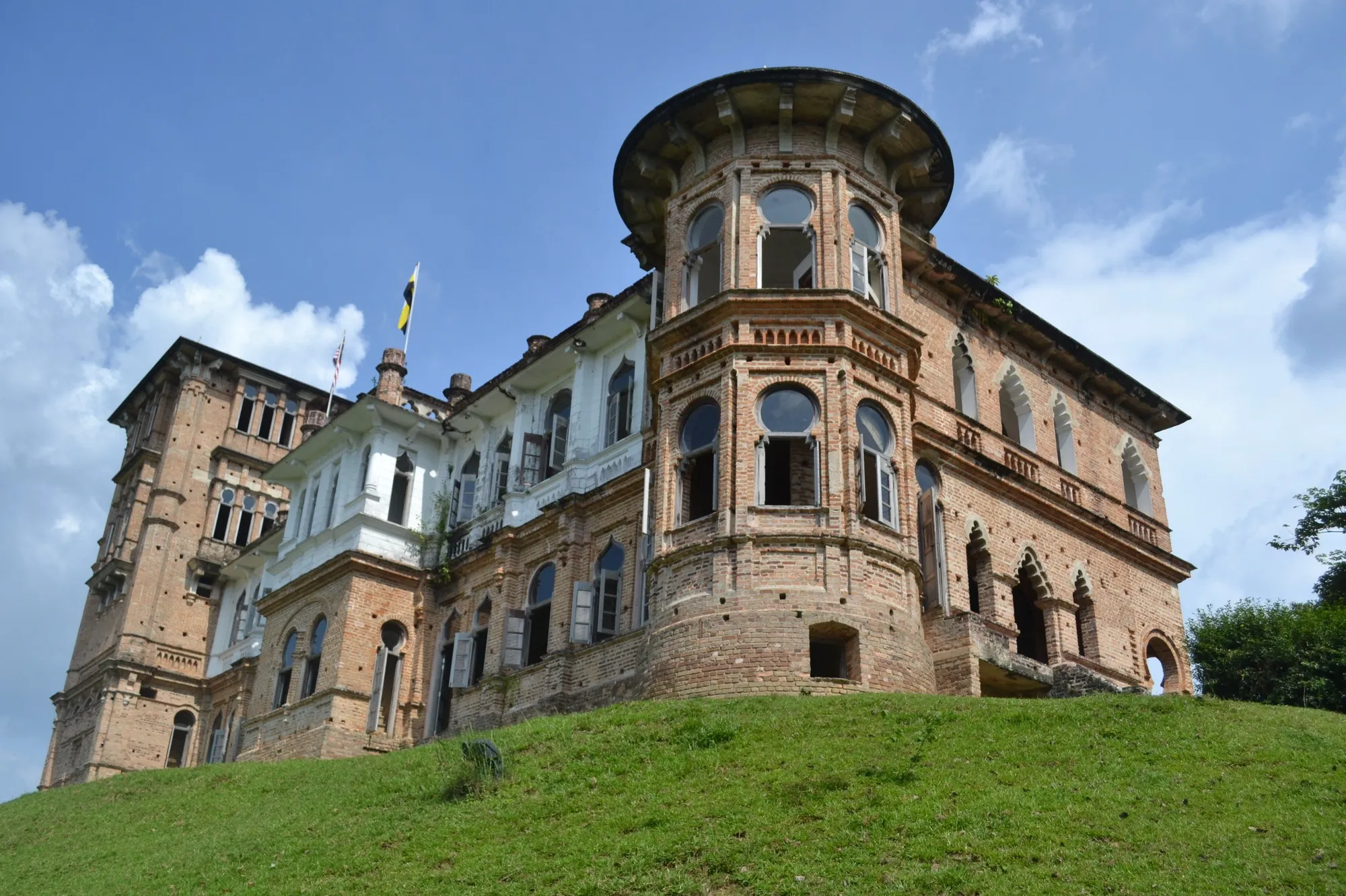 Unfinished red-brick mansion with a rounded tower, arched windows and a whitewashed wing on a grassy slope under a blue sky