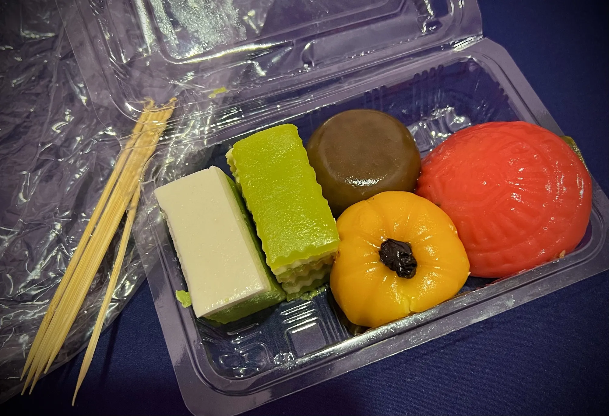 Box of colourful Nyonya kuih: layered green and yellow cakes, a brown round kuih and an orange-red ang ku kuih