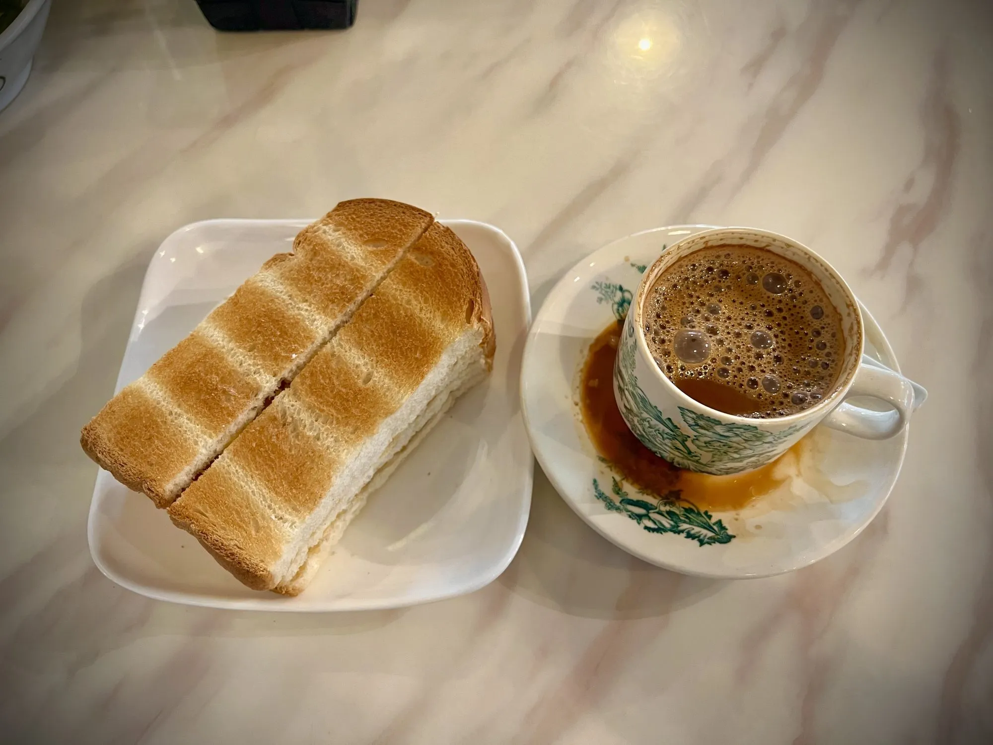 Hot frothy white coffee in a green floral cup and saucer beside toast on a marble table in Ipoh